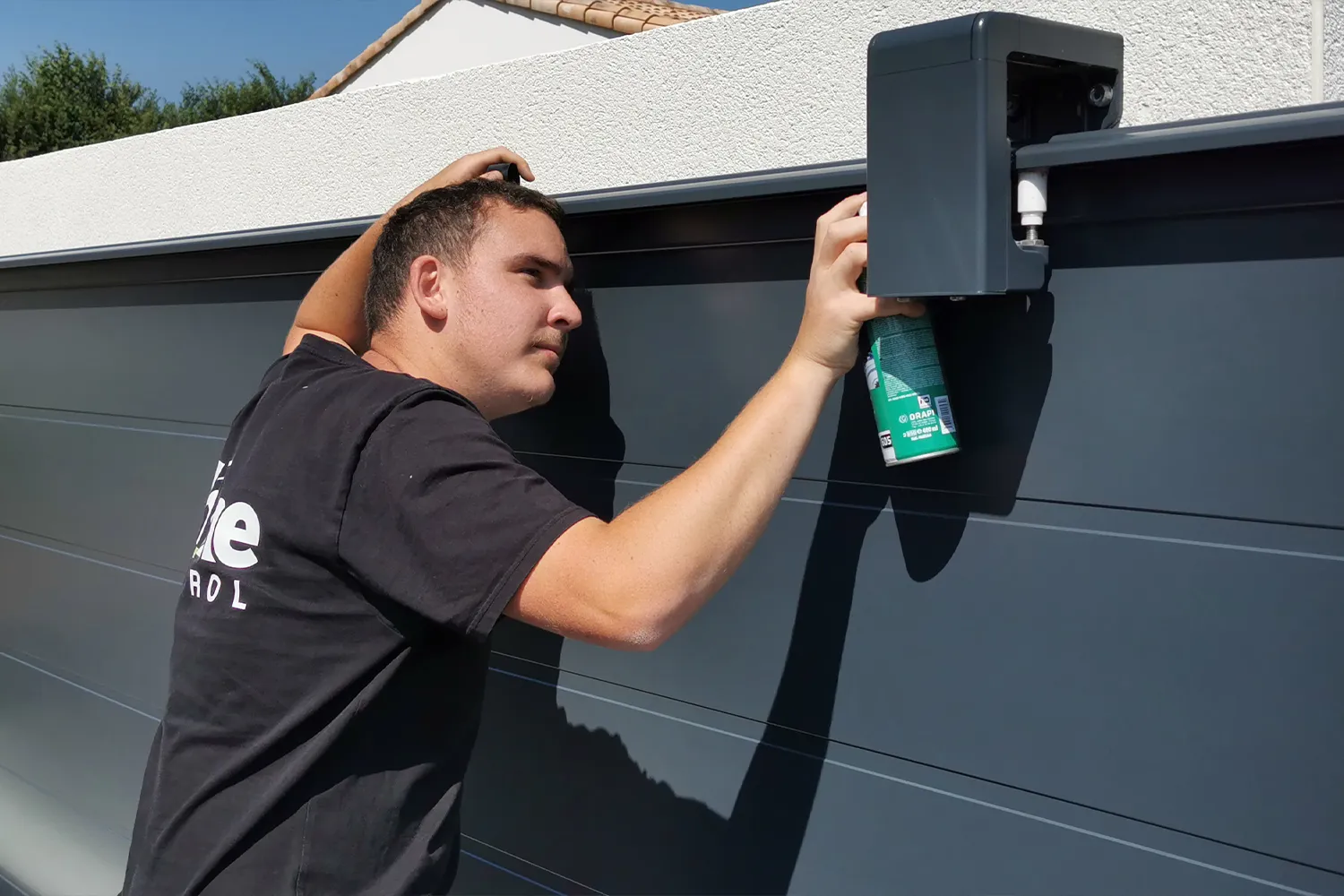 Technicien Home Control appliquant un lubrifiant sur les galets du guide haut d'un portail coulissant gris anthracite